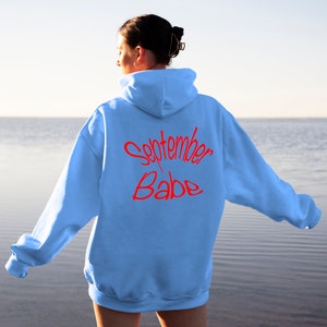 Puede incluir: Una sudadera de color azul claro con el texto "September Babe" en letras rojas en la parte posterior.