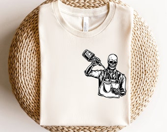 Puede incluir: Una camiseta blanca con una ilustración en blanco y negro de un esqueleto vertiendo una bebida de una botella. El esqueleto lleva un delantal y sostiene un vaso.