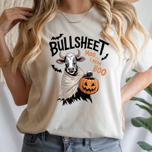 Peut inclure: T-shirt crème avec un motif sur le thème d'Halloween. Le motif comprend une vache déguisée en fantôme tenant une citrouille sculptée, avec les mots "BULLSHEET Moo... I MEAN BOO" et des illustrations de chauves-souris.