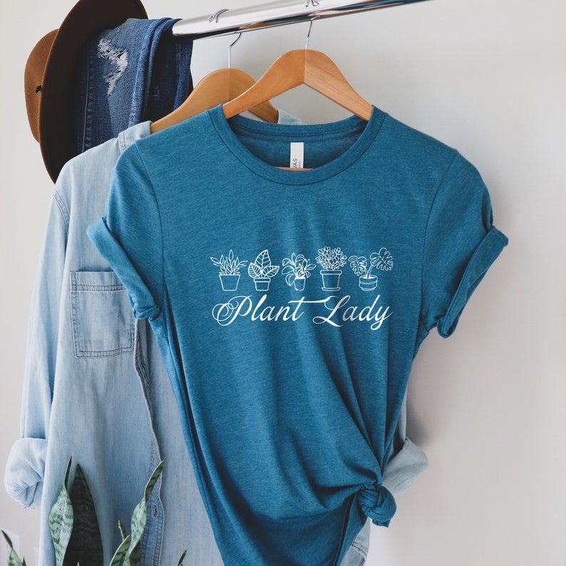 K&ouml;nnte beinhalten: Ein blaues T-Shirt mit einem wei&szlig;en Aufdruck von Topfpflanzen und dem Text "Plant Lady".