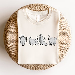 May include: A white t-shirt with a graphic of four cats dressed as ghosts. The cats are in a line, facing forward, and each has a ghost sheet draped over their body. The graphic is in black and white.