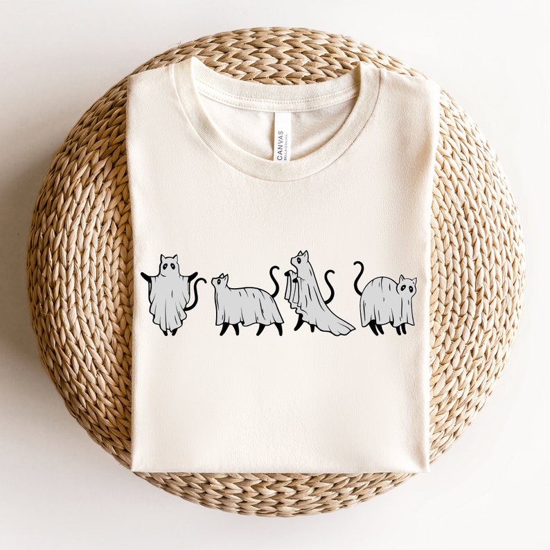 May include: A white t-shirt with a graphic of four cats dressed as ghosts. The cats are in a line, facing forward, and each has a ghost sheet draped over their body. The graphic is in black and white.