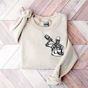 May include: A beige sweatshirt with a black and white graphic of a skeleton pouring a drink from a bottle into a glass. The graphic is centered on the front of the sweatshirt.