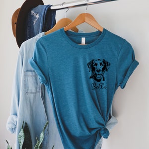 May include: A teal blue t-shirt with a black silhouette of a Labrador Retriever and the name "Bella" printed on the front.