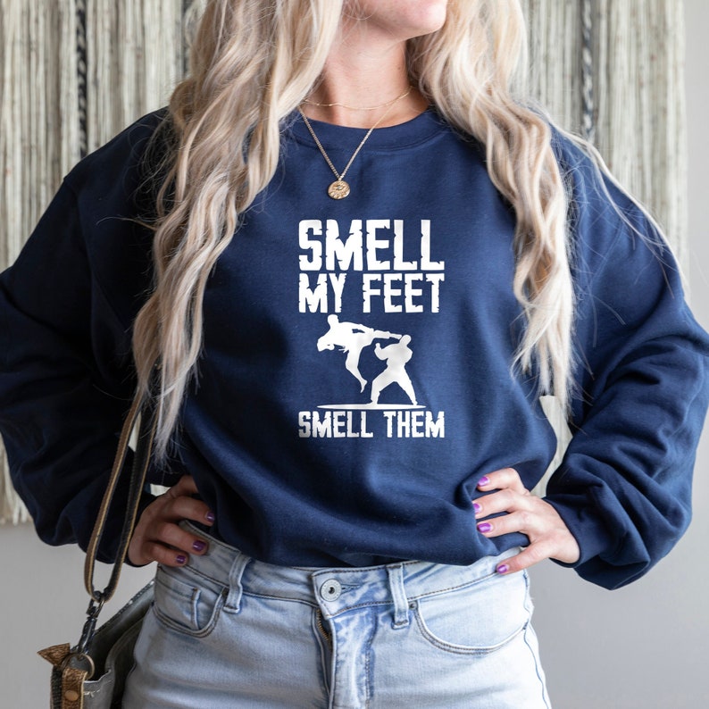Może przedstawiać: Bordowy sweter z napisem "Smell My Feet" i "Smell Them" w kolorze białym. Tekst jest otoczony grafiką przedstawiającą dwie osoby w pozie walki sztuk walki.