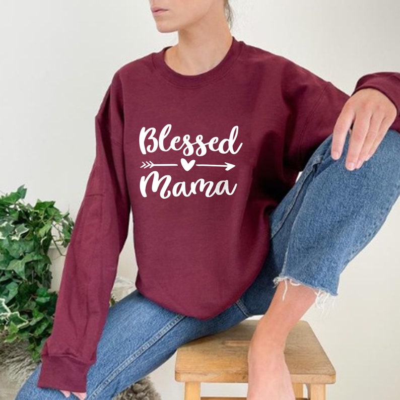 Puede incluir: Una sudadera color burdeos con el texto "Blessed Mama" en letras blancas con un peque&ntilde;o coraz&oacute;n y una flecha gr&aacute;fica.