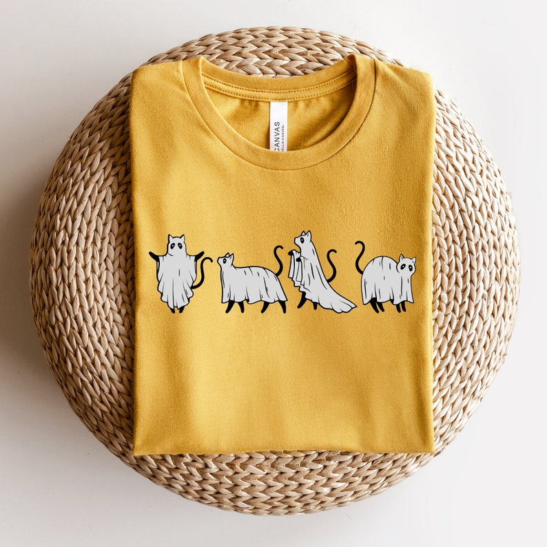 May include: A yellow t-shirt with a graphic of four cats dressed as ghosts. The cats are in a line, facing forward, and each has a white ghost sheet draped over their body. The text "Canvas" is visible on the shirt's tag.