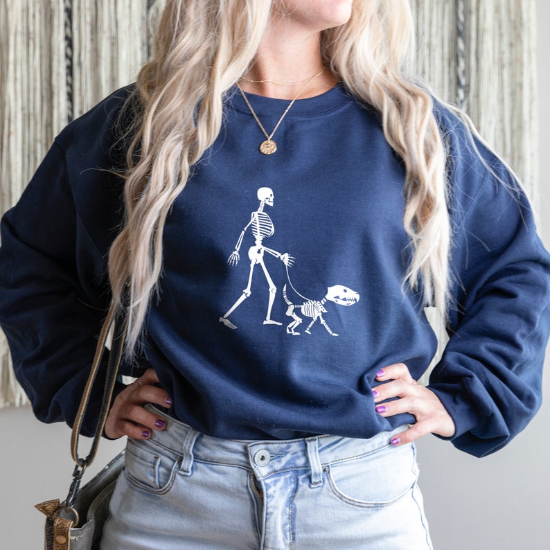 Pode incluir: Uma camisa de moletom azul marinho com um gr&aacute;fico branco de um esqueleto passeando um esqueleto de cachorro. A camisa de moletom tem gola redonda e mangas compridas.