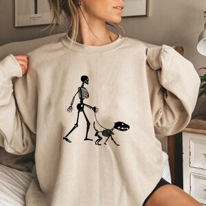 Pode incluir: Uma camisa de moletom bege com um gr&aacute;fico de um esqueleto passeando um cachorro esqueleto. O gr&aacute;fico &eacute; preto.