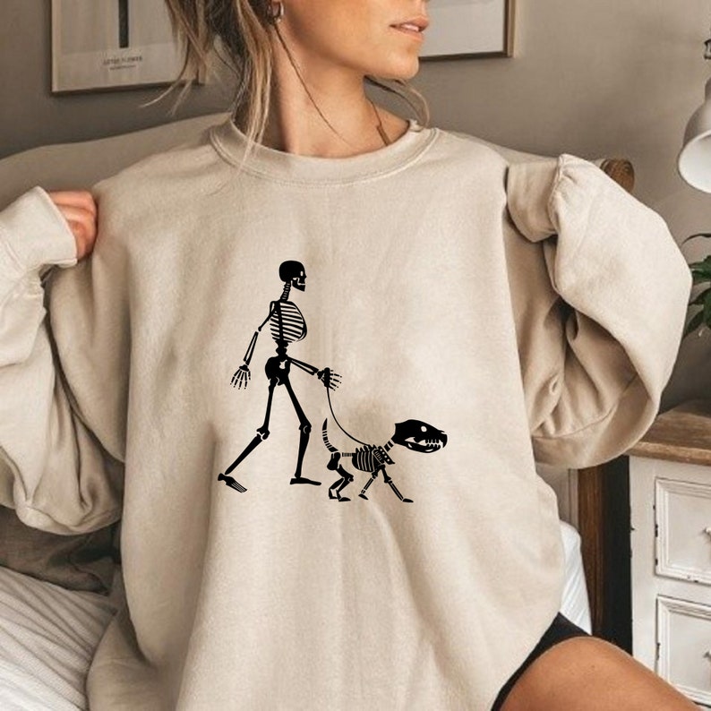 Pode incluir: Uma camisa de moletom bege com um gr&aacute;fico de um esqueleto passeando um cachorro esqueleto. O gr&aacute;fico &eacute; preto.
