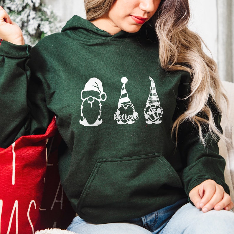May include: A dark green hooded sweatshirt with a white graphic of three gnomes wearing Santa hats. The middle gnome has the word "Believe" written below it.