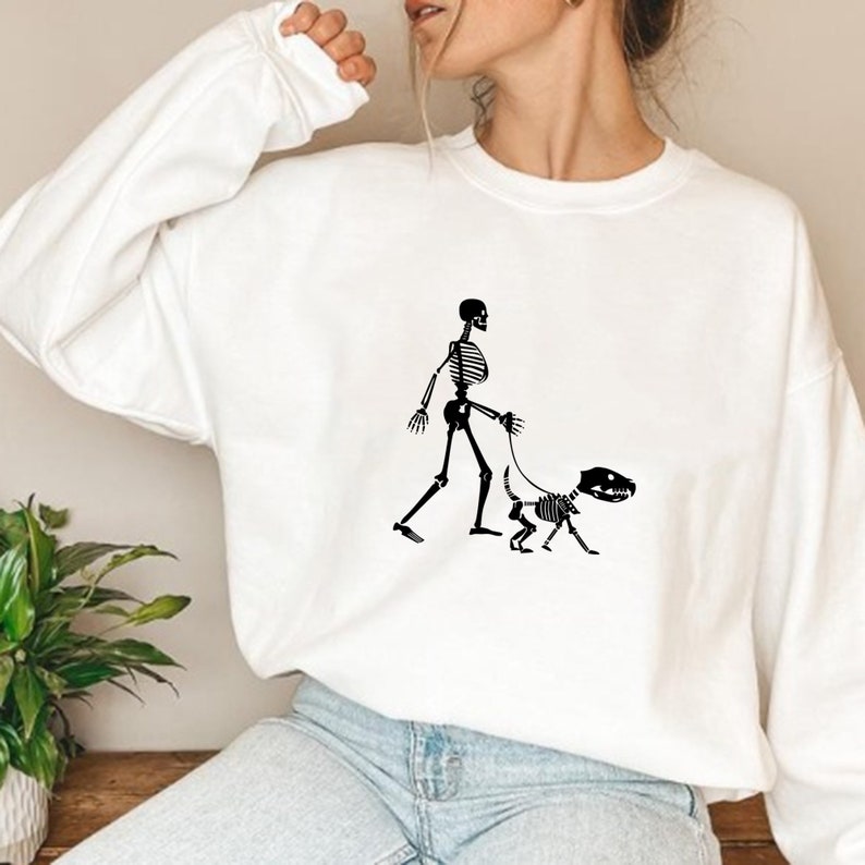 Pode incluir: Camisola branca com um gr&aacute;fico de esqueleto preto a passear um c&atilde;o esqueleto.