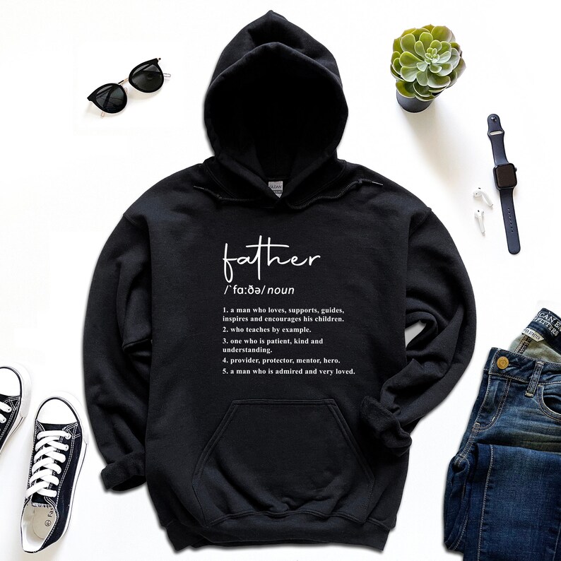 K&ouml;nnte beinhalten: Schwarzer Kapuzenpullover mit dem Wort "father" in wei&szlig;er Schrift. Unter dem Wort befindet sich eine Definition des Wortes "father". Der Pullover liegt auf einer wei&szlig;en Oberfl&auml;che mit Sonnenbrille, Uhr und Jeans.