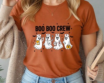 May include: An orange t-shirt with the text "BOO BOO CREW" above a graphic of four ghost characters dressed as medical professionals. Each ghost holds a different medical tool or supply, with bats and crosses around the design.