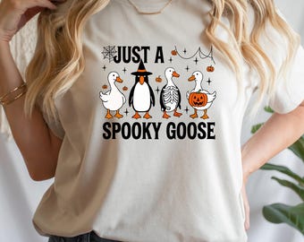 May include: A beige t-shirt with a Halloween design. The graphic shows four cartoon geese in spooky costumes, with the text "JUST A SPOOKY GOOSE". The geese are dressed as a witch, skeleton, and a pumpkin. The design includes cobwebs and small pumpkins.
