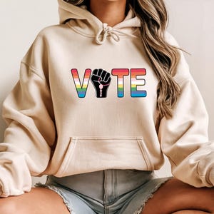 May include: A beige hoodie with a front pocket and a drawstring hood. The word "VOTE" is printed across the chest in rainbow colors, with a black fist graphic replacing the "O". The hoodie is made of a soft material.
