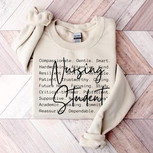Puede incluir: Una sudadera color crema con las palabras "Nursing Student" en fuente cursiva, con una lista de atributos positivos en negro, fuente sans-serif. La sudadera está doblada sobre una superficie de madera marrón claro.