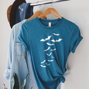 Puede incluir: Una camiseta azul turquesa con un gr&aacute;fico blanco de murci&eacute;lagos volando en bandada. La camiseta est&aacute; colgada en una percha de madera.