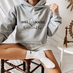 Może przedstawiać: Szara bluza z kapturem z napisem "anti social butterfly" i stylizowanym wzorem motyla.