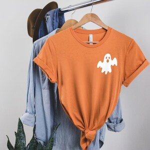 以下が含まれることがあります： オレンジ色の半袖Tシャツで、前面に白い幽霊のグラフィックが描かれています。幽霊は驚いた表情をしています。