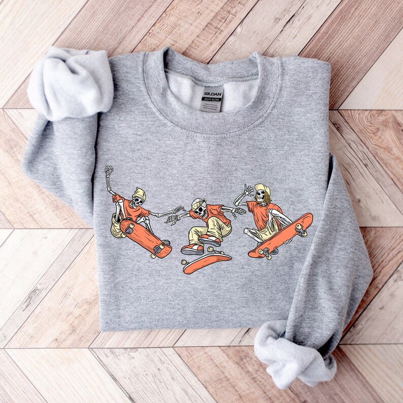 May include: Gray sweatshirt with a graphic of three skeletons skateboarding. The skeletons are wearing orange shirts and hats. The graphic is in a vintage style.