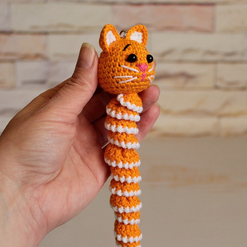 May include: A crocheted orange and white cat keychain with a curled tail. The cat has black eyes and a pink nose.