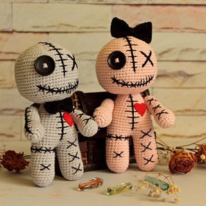 May include: Two handmade crochet dolls, one light gray and one light pink, featuring black button eyes, stitched X-shaped eyes, black stitched mouths, and red heart patches on their chests. The gray doll wears a black bow tie, and the pink doll has a black bow.
