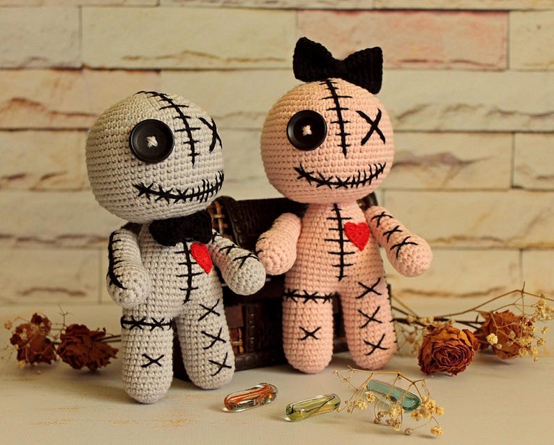 May include: Two handmade crochet dolls, one light gray and one light pink, featuring black button eyes, stitched X-shaped eyes, black stitched mouths, and red heart patches on their chests. The gray doll wears a black bow tie, and the pink doll has a black bow.