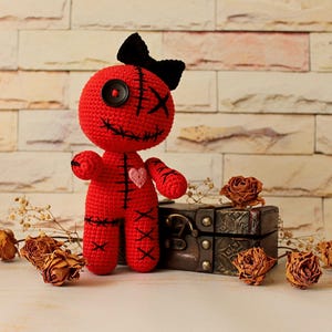 May include: A red crocheted doll featuring a black button eye, a black stitched eye, and a black stitched mouth. The doll has a black bow on its head, a small pink heart on its chest, and black stitched details on its arms, legs, and torso.