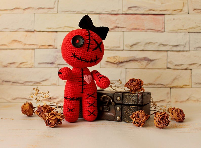 May include: A red crocheted doll featuring a black button eye, a black stitched eye, and a black stitched mouth. The doll has a black bow on its head, a small pink heart on its chest, and black stitched details on its arms, legs, and torso.