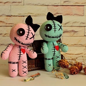 May include: Two handmade crochet dolls, one light pink and one mint green, each featuring a black yarn bow on top, a single black button eye, a stitched X eye, and a stitched mouth. Both dolls have a small red heart patch on the chest.