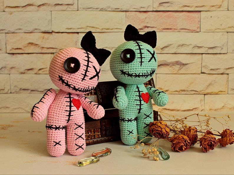 May include: Two handmade crochet dolls, one light pink and one mint green, each featuring a black yarn bow on top, a single black button eye, a stitched X eye, and a stitched mouth. Both dolls have a small red heart patch on the chest.