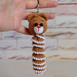 May include: A brown crocheted bear keychain with a white crocheted tail. The bear has black eyes and a red nose.