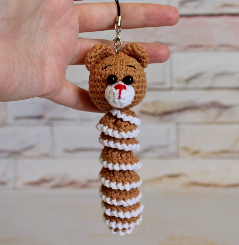 May include: A brown crocheted bear keychain with a white crocheted tail. The bear has black eyes and a red nose.