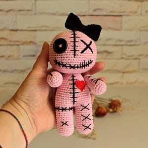 May include: A light pink crochet doll featuring a black bow on its head, one black button eye, one black stitched X eye, and a stitched mouth. The doll has a red heart on its chest and black stitched details on its body, arms, and legs.