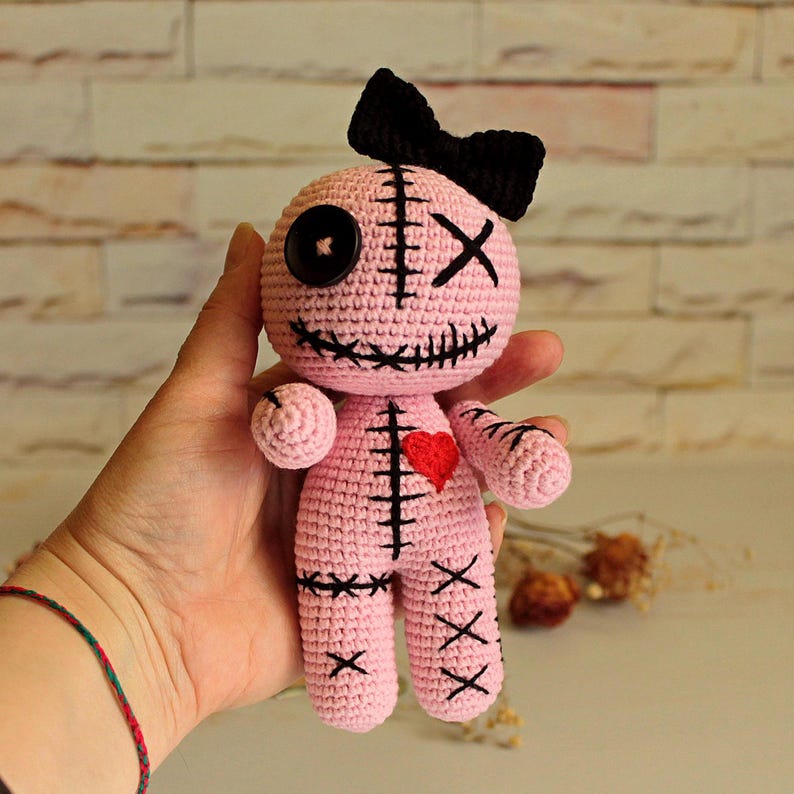 May include: A light pink crochet doll featuring a black bow on its head, one black button eye, one black stitched X eye, and a stitched mouth. The doll has a red heart on its chest and black stitched details on its body, arms, and legs.