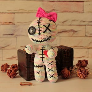 May include: A handmade crochet doll featuring a pastel variegated yarn body with black stitched details, a single black button eye, an X-shaped eye, a stitched mouth, and a bright pink bow on top. A small red heart is stitched onto the chest.
