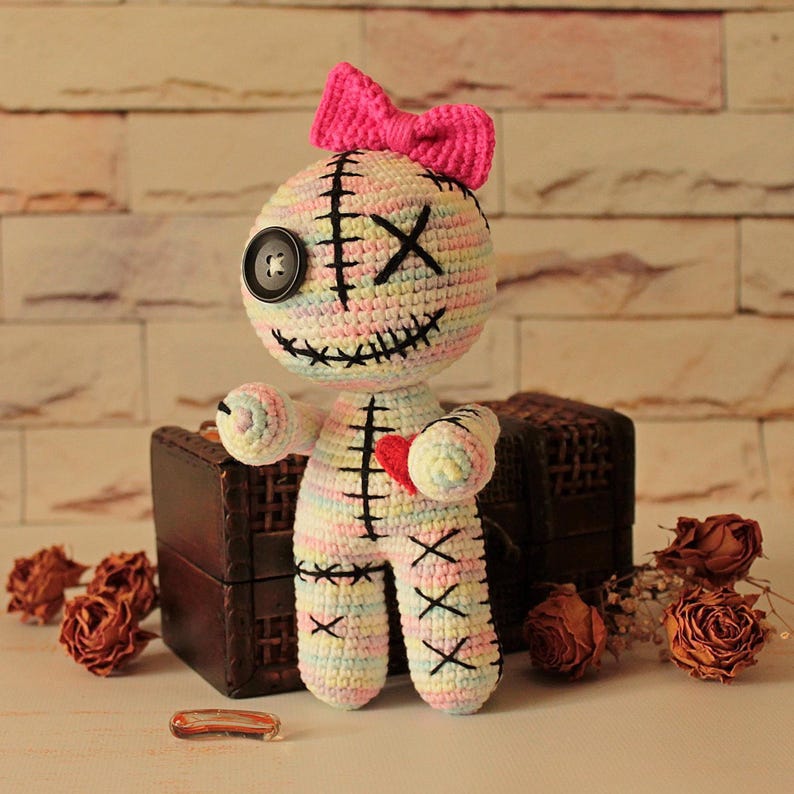 May include: A handmade crochet doll featuring a pastel variegated yarn body with black stitched details, a single black button eye, an X-shaped eye, a stitched mouth, and a bright pink bow on top. A small red heart is stitched onto the chest.