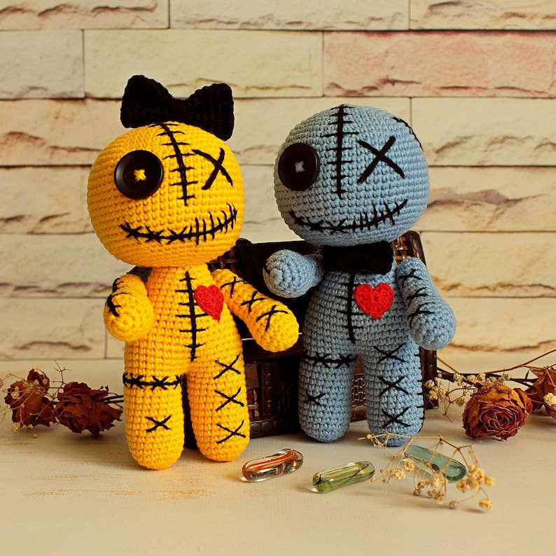 May include: Two handmade crochet dolls, one yellow and one light blue, featuring black button eyes, stitched X-shaped eyes, and embroidered black mouths. Each doll has a small red heart on its chest and black stitched details on the limbs.