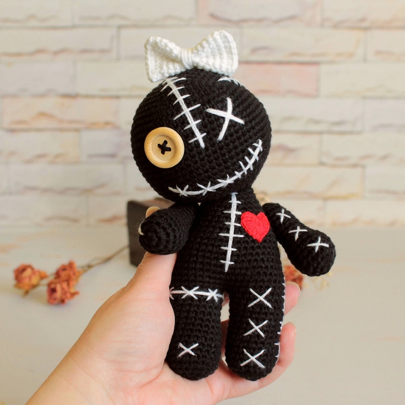 May include: A black crocheted doll featuring a white bow on its head, a beige button for one eye, and white stitched X-shaped details across its body. A small red heart is stitched onto the chest area, and the doll has a round head and limbs.