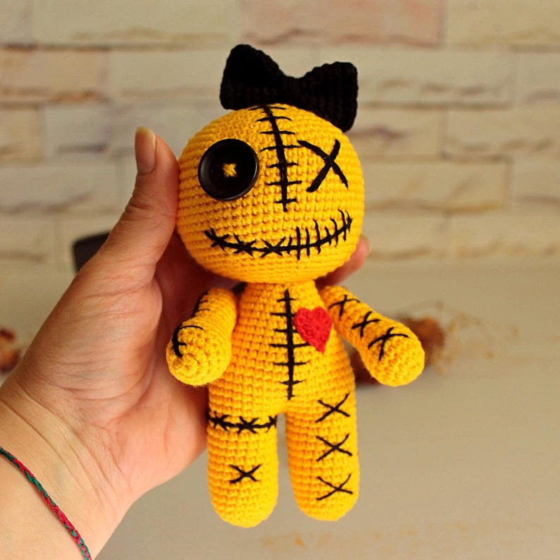 May include: A yellow crocheted doll featuring a black bow on its head, one black button eye, one stitched X eye, and a stitched mouth. The doll has a red heart patch on its chest and black stitched X patterns on its arms, legs, and torso.