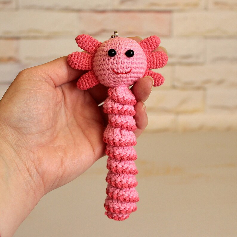 May include: Pink crocheted axolotl keychain with black eyes and a smiling mouth. The axolotl is holding a small silver ring.