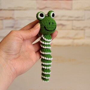 May include: Green crocheted frog keychain with white accents. The frog has large black eyes and a smiling mouth. The tail is long and segmented.