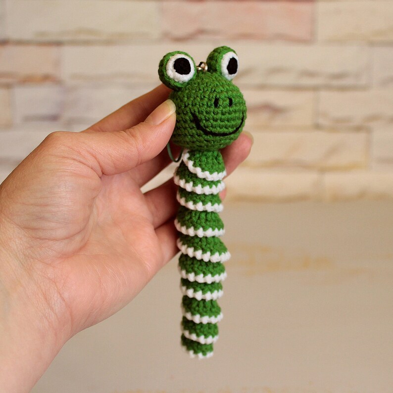 May include: Green crocheted frog keychain with white accents. The frog has large black eyes and a smiling mouth. The tail is long and segmented.