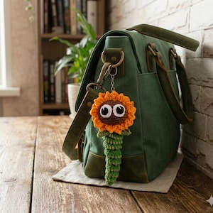 May include: A dark green canvas messenger bag with leather trim and a metal clasp. Attached to the clasp is a crocheted sunflower keychain featuring a brown center, bright orange petals, large white and black googly eyes, and a green stem.