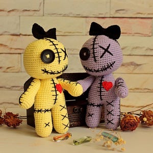May include: Two handmade crochet dolls, one yellow and one light purple, each featuring a black button eye, an X-shaped eye, a stitched mouth, a black bow on top, and a red heart patch on the chest, with black stitched details on the limbs and torso.