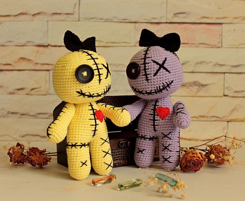 May include: Two handmade crochet dolls, one yellow and one light purple, each featuring a black button eye, an X-shaped eye, a stitched mouth, a black bow on top, and a red heart patch on the chest, with black stitched details on the limbs and torso.