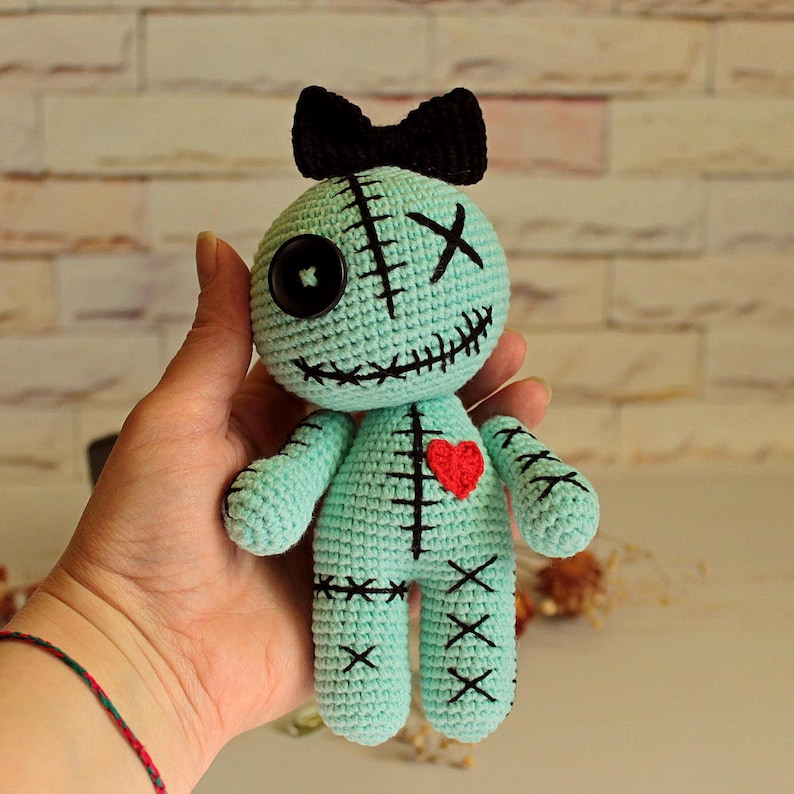 May include: A light mint green crochet doll featuring a black bow on its head, one black button eye, one stitched X eye, and a stitched mouth. The doll has a red heart embroidered on its chest and black stitched details across its body.