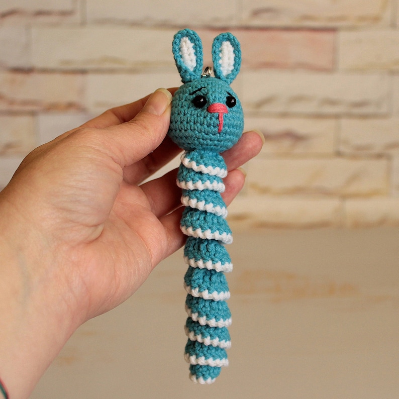 May include: A crocheted blue and white bunny keychain with a long, coiled tail. The bunny has a pink nose and black eyes.