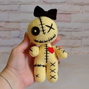 May include: A cream-colored crochet doll featuring a black yarn bow on its head, one large black button eye, one stitched X eye, and a stitched mouth. The body has black cross-stitch detailing and a small red heart patch on the upper chest area.